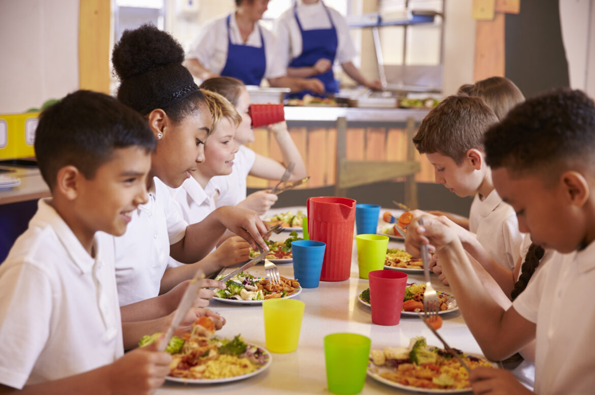 Elementary school kids gather around a cafeteria lunch table with plates full of food. They are socializing with each other and sharing a meal.